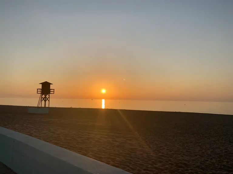 An empty lifeguard watchtower silhouetted against the sunrise on Playa de Levante in La Linea de la Concepcion, with flat sea and empty sand.